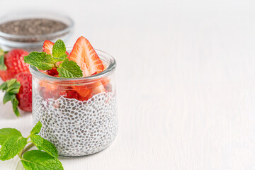 Sweet chia pudding dessert made with seeds and plant based milk with sliced juicy ripe strawberries topping and fresh mint leaf served in glass jar on white wooden table with copy space and ingredient