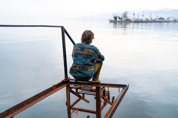 Pensive man sitting alone on weathered pier by tranquil sea at dusk, horizon stretching beyond.