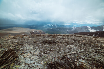 Dramatic panoramic view from high stony pass to alpine valley against large snow-capped mountain range in rainy low clouds. Sharp stones on hill against misty snowy mountains in rain under cloudy sky.
