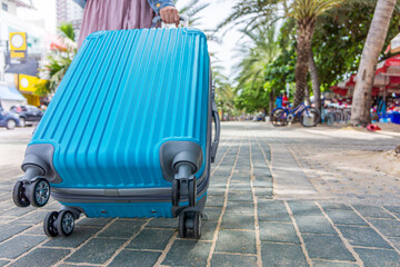 A female tourist is dragging a blue wheeled luggage on the sidewalk. She is traveling to various tourist attractions in the city.