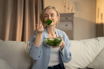 A joyful woman is seated comfortably on a couch, holding a bowl of fresh green salad. She appears...