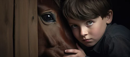 Tuinposter A young boy is standing next to a horse, peering out from behind the ponys body. The boy appears curious and cautious, while the horse stands calmly. © TheWaterMeloonProjec