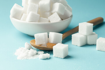 Sugar cube in wood spoon, closeup with bowl full of sugar on blue background