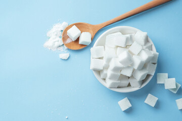 Pile of sugar cubes in white bowl with wood spoon on light blue background, top view