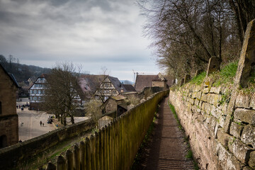 Ein mittelalterlicher Pfad, der mit einer Mauer und einem Holzzaun gesäumt ist, verläuft oberhalb eines Dorfplatzes mit alten Fachwerkhäusern