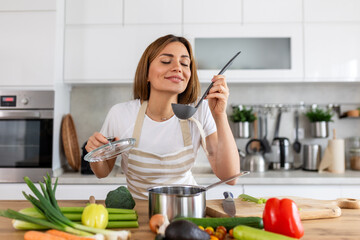 Happy Young Woman Cooking Tasting Dinner In A Pot Standing In Modern Kitchen At Home. Housewife...