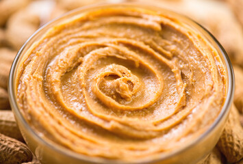 Peanut butter swirls in a glass bowl over raw peanuts background. Creamy smooth peanut butter in jar backdrop, organic food. American cuisine. - 755417744