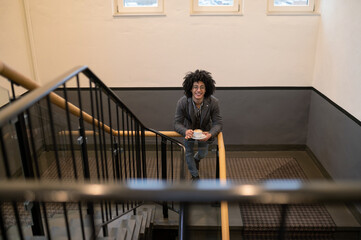 Curly-haired young man in eyeglasses at the stairs with a cup in hands