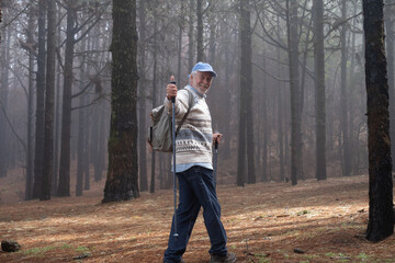 Active happy senior retired man with backpack walking in mountain forest on a foggy day with the help of poles enjoying nature, freedom and free time. Forest background with bare trees