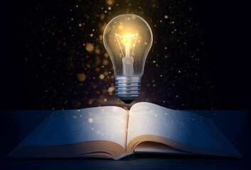 Glowing light bulb over a textbook on a dark background with glitter.  Concept to produce new idea based on knowledge. - obrazy, fototapety, plakaty