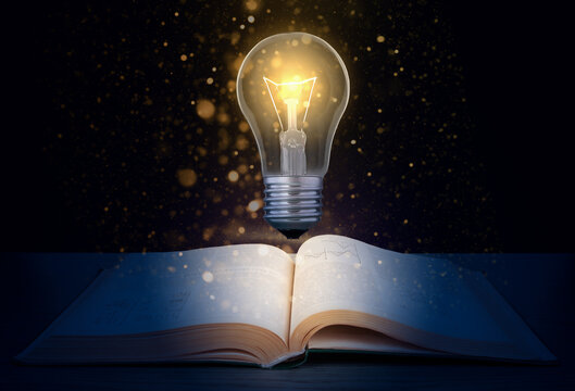 Glowing light bulb over a textbook on a dark background with glitter.  Concept to produce new idea based on knowledge.