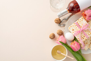 Overhead shot featuring a beautifully arranged Passover plate with matzah wrapped in ribbon, wine bottle, glass, nuts with a nutcracker, honey, a dipper, an egg, and tulips, all set on beige backdrop