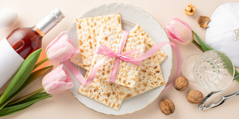 Top-view image showcasing a Passover seder setup with ribbon-tied matzah, red wine, and a glass, alongside nuts, a nutcracker, kippah, an egg, and tulips on a soft beige background