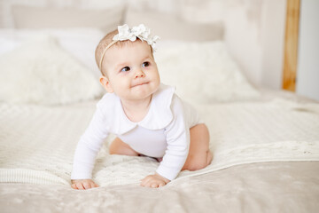 portrait of a little baby girl of six months on a bed at home in a bright bedroom smiling, a happy newborn in a white cotton bodysuit crawls and looks away