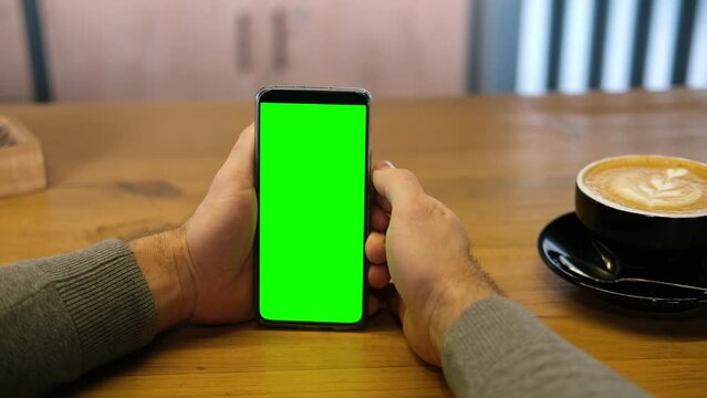 Young man sitting at cafe holding smartphone green mock-up screen in hand. Male person using chroma key mobile phone. Vertical mode. Touching, swiping display, tapping, surfing internet social media	