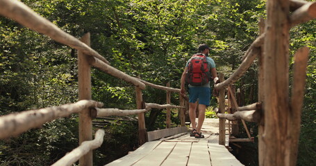A man in hiking gear with a backpack walks across an epic suspension bridge in a national park. A cinematic adventure of traveling in the mountains. A man walks through a small town on a mountain rive