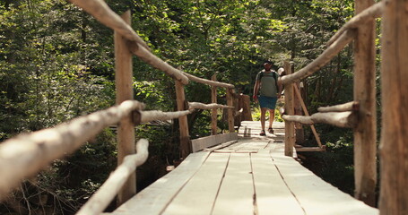 A man in hiking gear with a backpack walks across an epic suspension bridge in a national park. A...