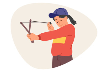 Little mischievous girl shoots with slingshot, closing one eye to aim at target during weekend. Child misbehaves by shooting at peers or glass with homemade slingshot with stretched rubber band.