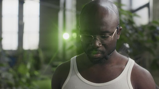 Tilting down shot of young Black man in tight white undershirt video calling on digital tablet with colleague while working at plant factory