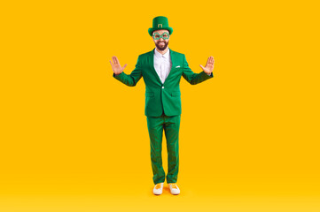 Portrait of a happy man in a leprechaun costume. Full body shot of a funny guy wearing a green suit, hat and shamrock party glasses standing on a yellow studio background. St Patrick's Day concept