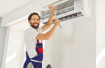 Happy male worker standing on ladder by white wall in the house, setting up new AC unit, fixing...
