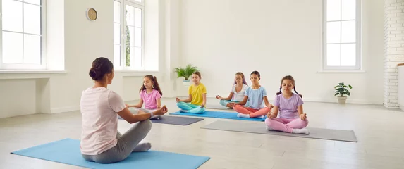 Fototapeten Children doing yoga with trainer. Kids doing relaxing exercises at gym. Group of little girls in sportswear sitting on rubber mats on floor in lotus pose and learning to mediate with woman coach © Studio Romantic