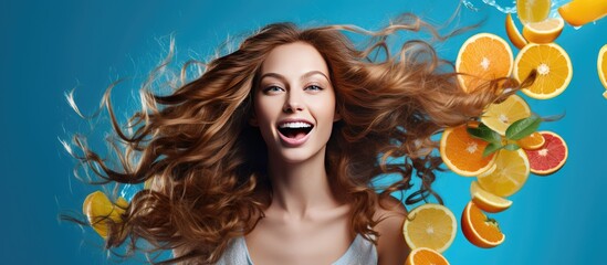 A happy young woman with long hair is surrounded by oranges, her hair flowing upwards. She wears a striped t-shirt and has citrus fruits on her head against a blue background. - obrazy, fototapety, plakaty