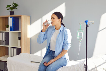 Young woman with sterile needle in arm sitting on medical bed by pole with IV drip bottle, drinking...