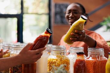 Storekeeper helping client looking for healthy pasta sauce in local neighborhood shop. Close-up of seller offering customer details about product in environmentally responsible zero waste store