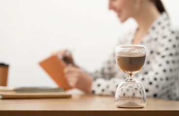 Hourglass with flowing sand on desk. Woman working indoors, selective focus
