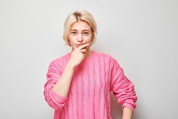 Thoughtful young woman in pink sweater with hand on chin looking at camera on light grey background. - obrazy, fototapety, plakaty
