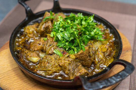 Close-up of the traditional Georgian dish ostri sprinkled with herbs in a cast-iron pan on a wooden board on the table.