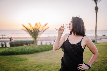 A woman in a black dress sips champagne by the seaside at sunset, with palm trees in the background and sunglasses atop her head