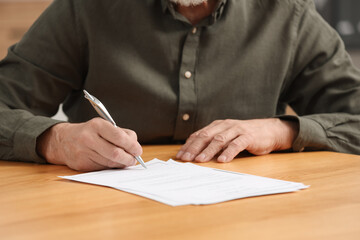 Senior man signing Last Will and Testament at wooden table indoors, closeup