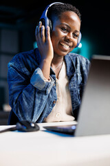 Black woman joyfully using a laptop and wireless headphones. African american female influencer...