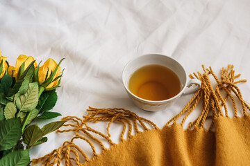 Eine Teetasse, ein gelber Rosenstrauss und eine Wolldecke mit Fransen auf einer weißen Bettdecke....