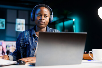 Focused black woman wearing wireless headphones using laptop and writing notes. African american female student with headset watching online webinar, listening to audio course on personal computer.