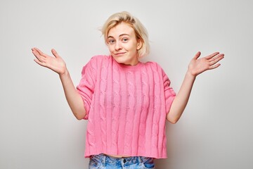 Woman in pink sweater shrugging with uncertain expression, isolated on grey background.