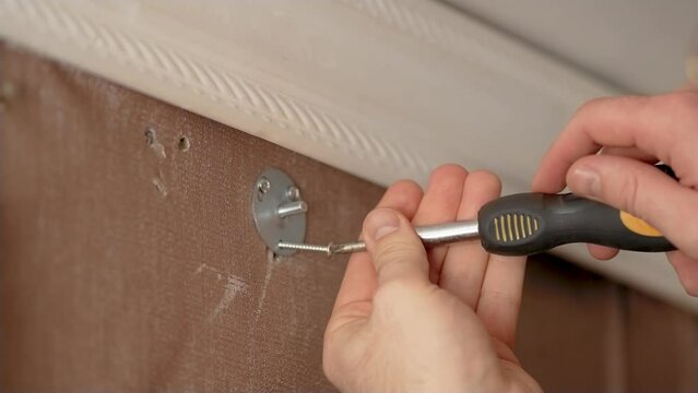 Close up man screws iron screw or bolt into previously drilled holes in wall to hang cornice in old apartment. Concept cheap home renovation. Create cheap comfort. Hands with screwdriver close up