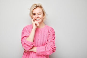 Smiling woman in pink sweater posing with hand on chin against a gray background.