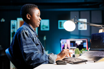 Fototapeta premium Female freelancer works at a desk, using a laptop to manage a futuristic information system. African american woman using her personal computer for cybersecurity and programming, working at home.