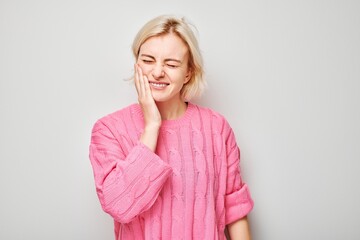 Smiling woman in pink sweater with hand on cheek against a gray background, expressing toothache.