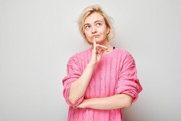 Thoughtful young woman in pink shirt looking up with finger on chin against a gray background.