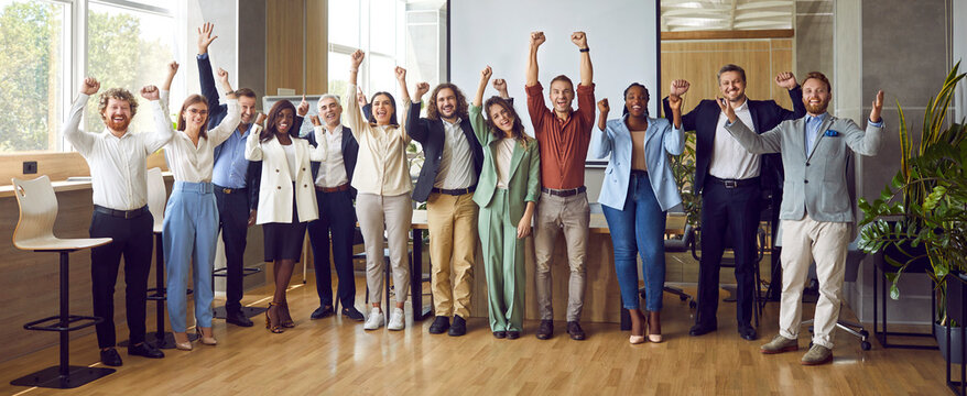 Group portrait of happy business team rejoicing in successful results of company. Smiling business men and women stand in row in office and joyfully clench their fists and shout in victory. Banner.