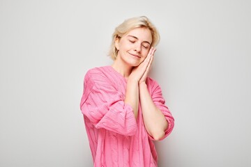 Woman with closed eyes smiling pretending to sleep with hands under cheek on gray background.