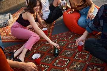 cheerful young multicultural friends in vivid attires playing spin the bottle at rooftop party