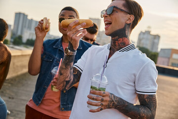 joyous multiracial friends in urban vivid outfits enjoying delicious hot dogs on rooftop at party
