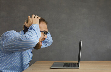 Side profile view man sitting at desk, holding head in hands, looking at laptop screen in despair. Male worker accidentally deletes important data or can't fix computer error and program malfunction