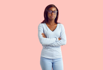 Portrait of serious female college or high school student. Young African American woman or teenage girl wearing blue long sleeve top, jeans and glasses standing, arms folded, on pastel pink background