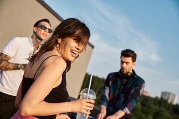focus on joyous attractive woman looking at camera next to her blurred friends at rooftop party